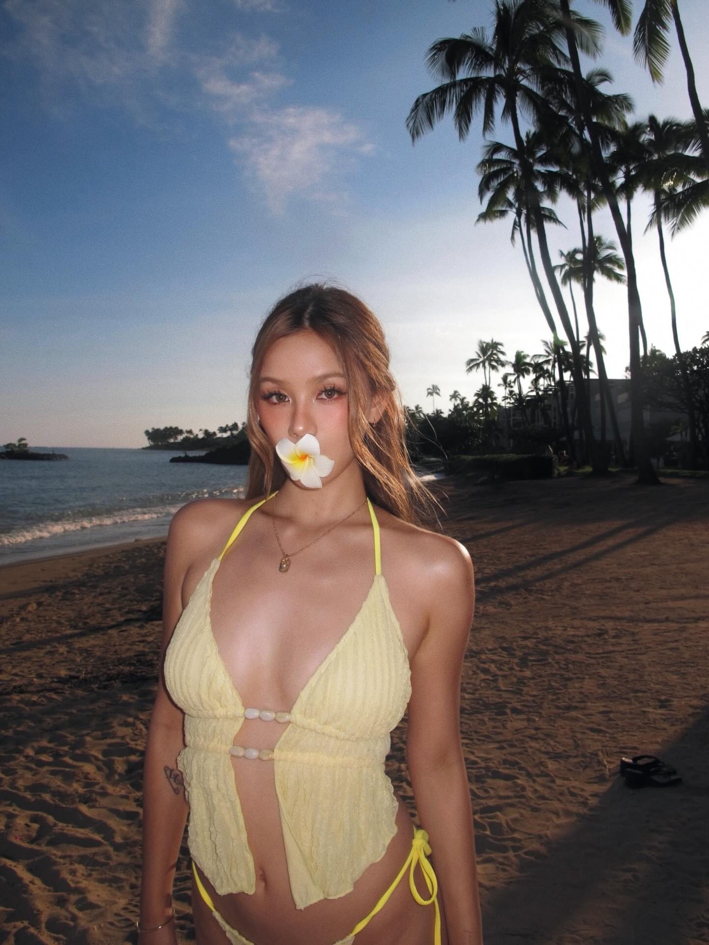 Kathy Zheng on a palm-lined beach holding a plumeria flower in her mouth