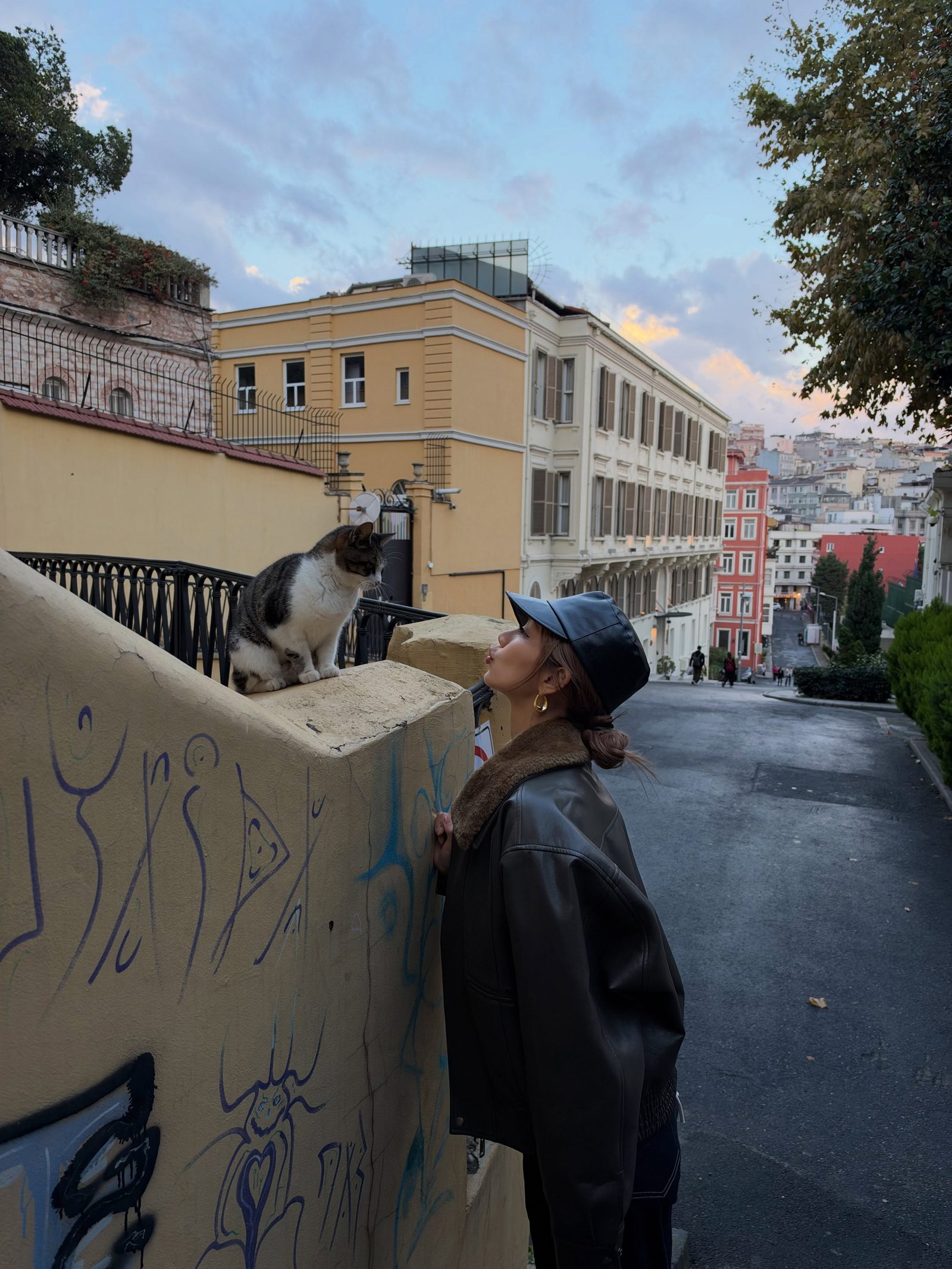 Kathy Zheng in a leather cap blowing a kiss to a cat on a wall