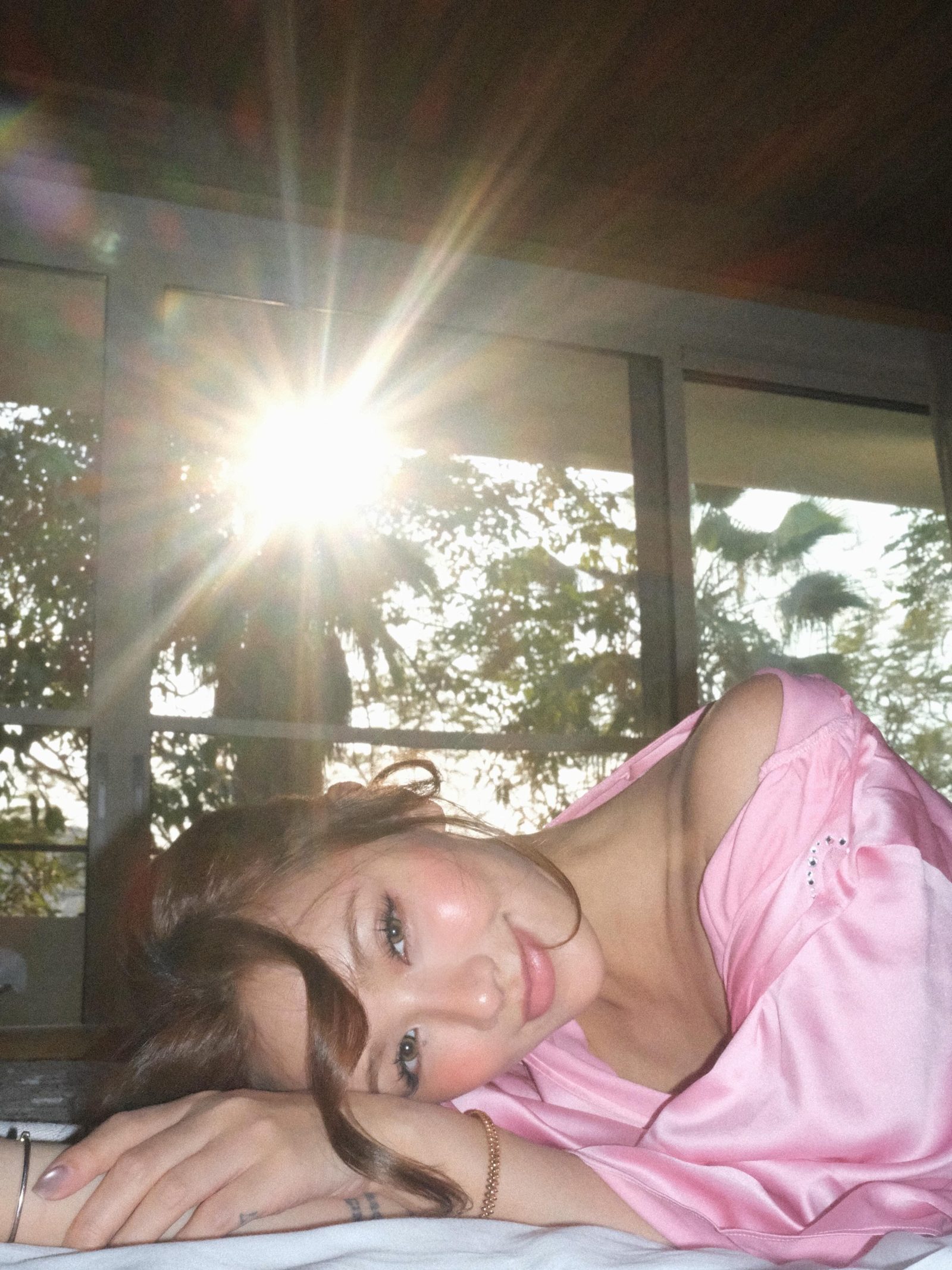 Kathy Zheng in pink satin lying by a window with a bright sun flare